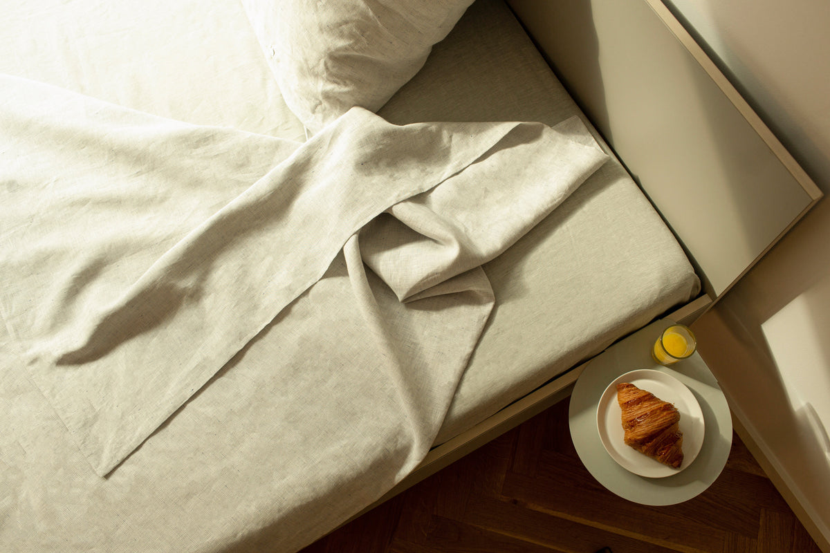 Bett mit Bettwäsche aus Leinen und einem Nachttisch daneben, auf dem Croissant und Saft stehen.