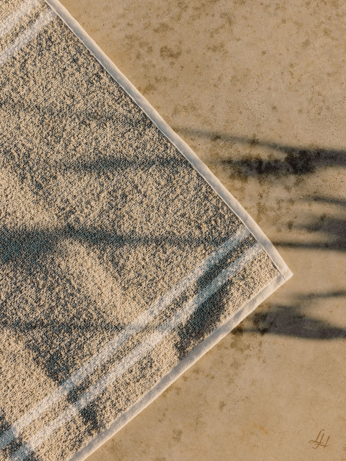 Ein beigefarbenes Handtuch mit weißen Streifen aus der Leinenmanufaktur liegt sonnenbeschienen auf Beton, durchsetzt mit Pflanzenschatten.