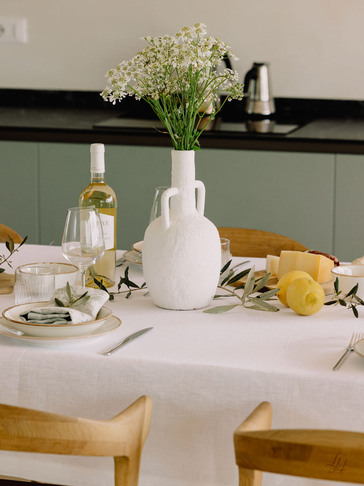 Eine weiße Vase mit Blumen steht auf einem gedeckten Esstisch mit Leinen Tischwäsche und Wein in einer modernen Küche.