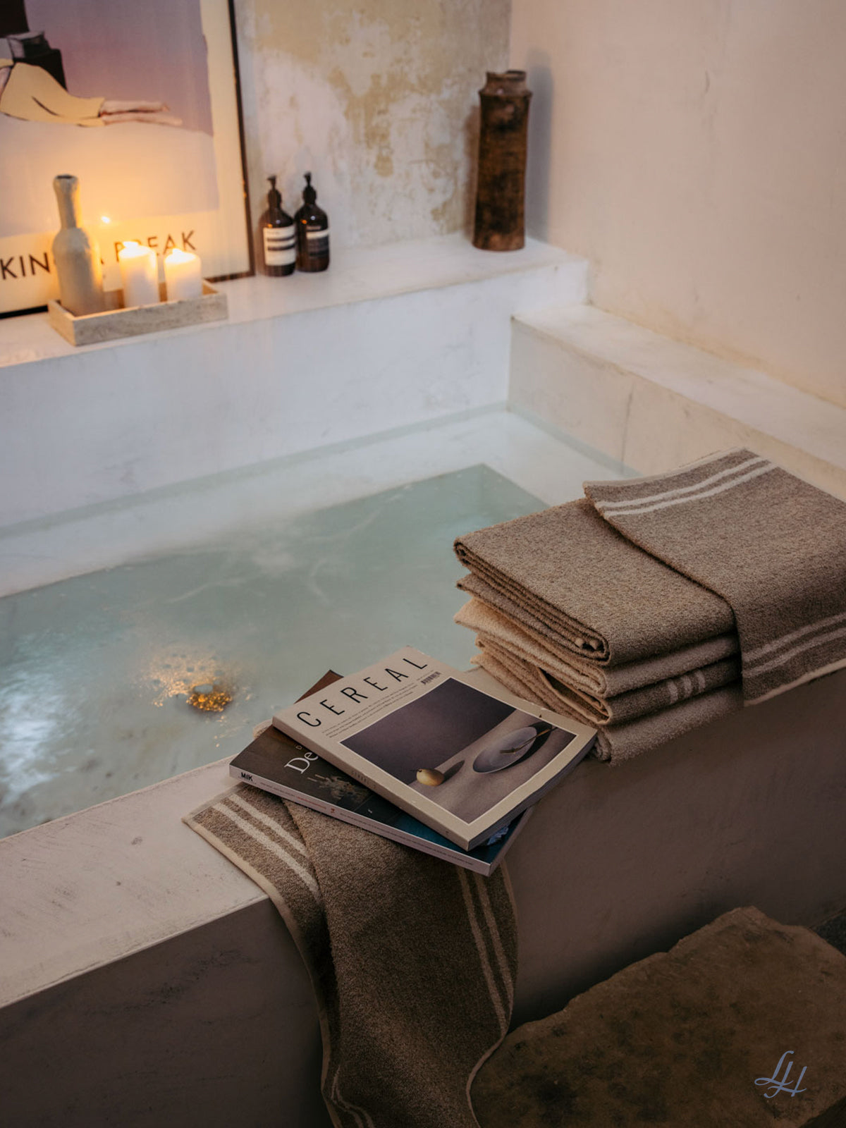 Eine gefüllte Badewanne mit Waschlappen aus Leinen, gefaltete Handtücher, Zeitschriften, Kerzen und Flaschen in einem sanft beleuchteten Raum.