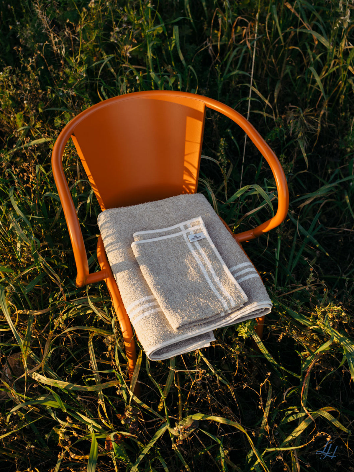 Ein orangefarbener Metallstuhl im hohen Gras hält gefaltete beige Leinenfrotté-Waschlappen aus Leinen auf seiner Sitzfläche.