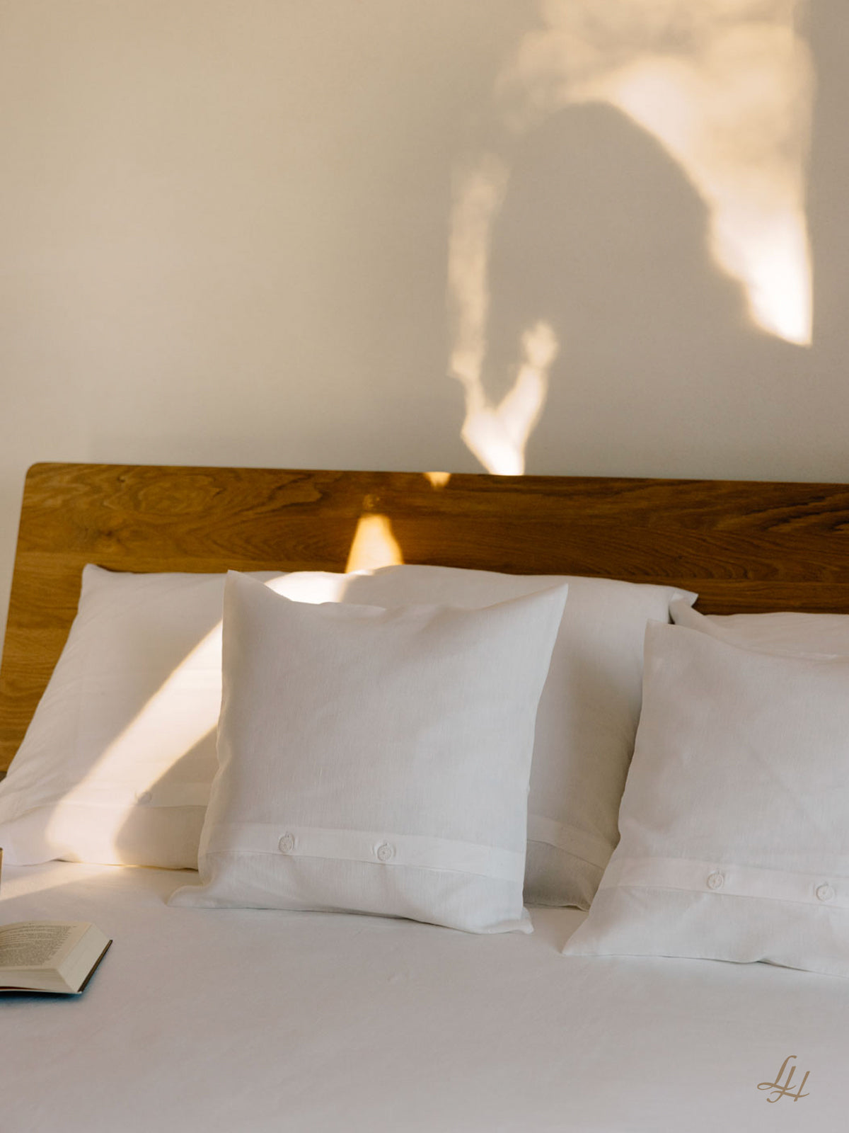 Ein ordentlich gemachtes Bett mit drei weißen Kissen in Leinenbettwäsche, ein aufgeschlagenes Buch und ein Schatten einer Person an der Wand hinter dem Kopfteil.