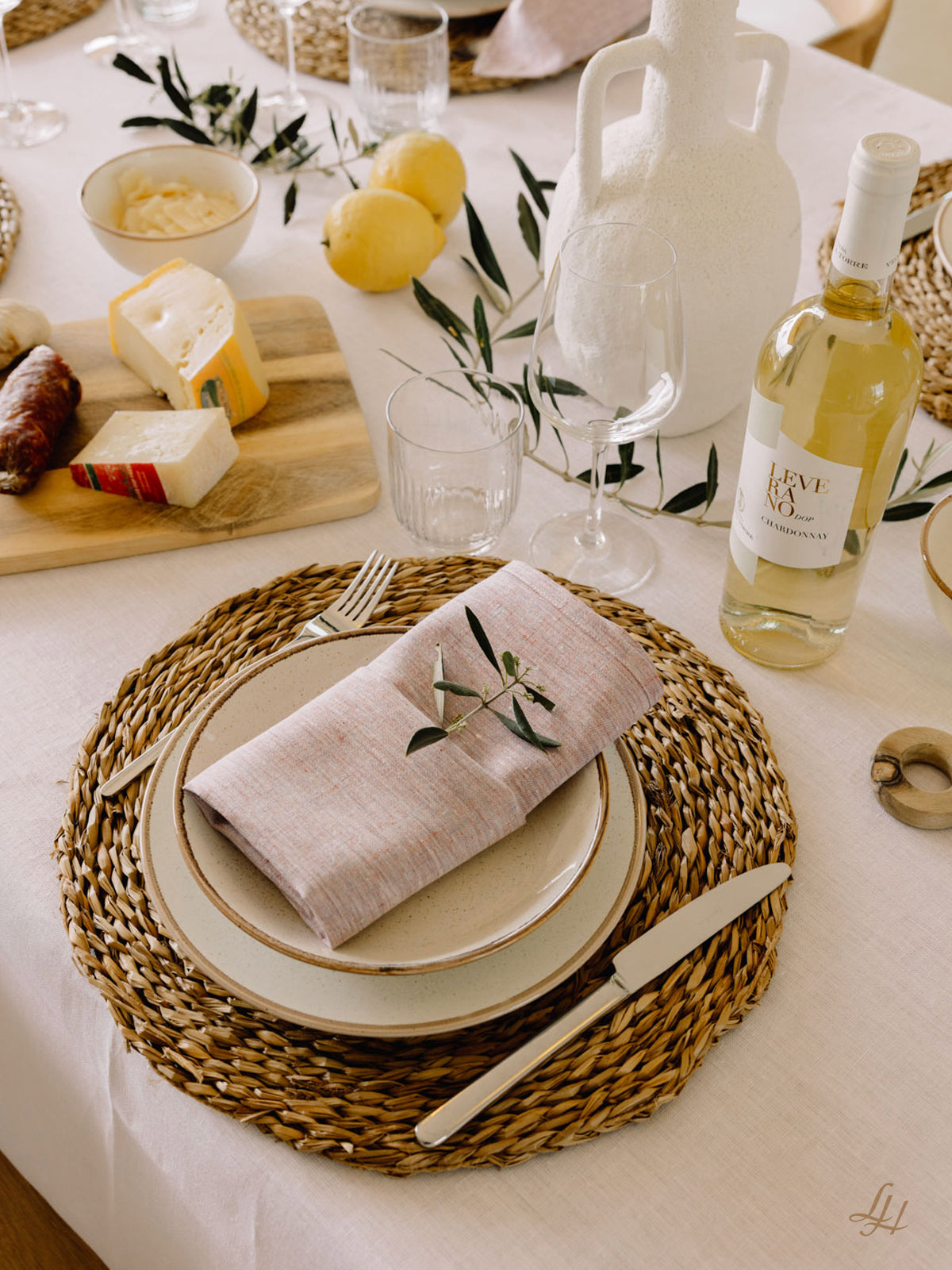 Ein gewebtes Tischset mit einem Teller, Besteck und hypoallergenen Leinenservietten auf einem mit Wein, Käse und Zitronen gedeckten Tisch.