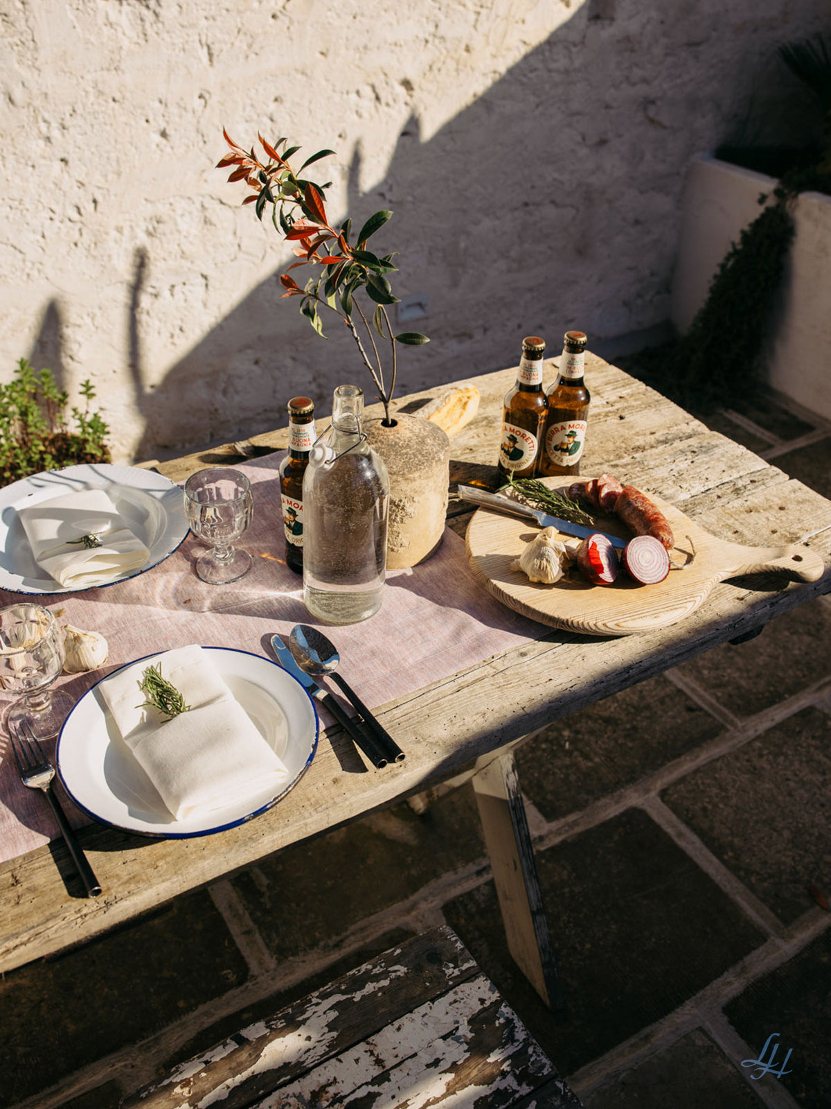 Ein rustikaler Outdoor-Tisch mit Tellern, Besteck, Bier, Brot und Wurst auf eleganter Leinen-Tischwäsche von Leinenmanufaktur.