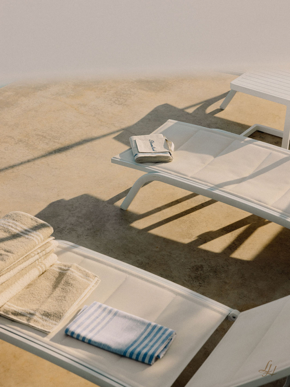 Zwei weiße Liegestühle mit Mangelleinen Strandtüchern stehen neben einem kleinen Tisch auf einer sonnenbeschienenen Terrasse mit weichen Schatten.