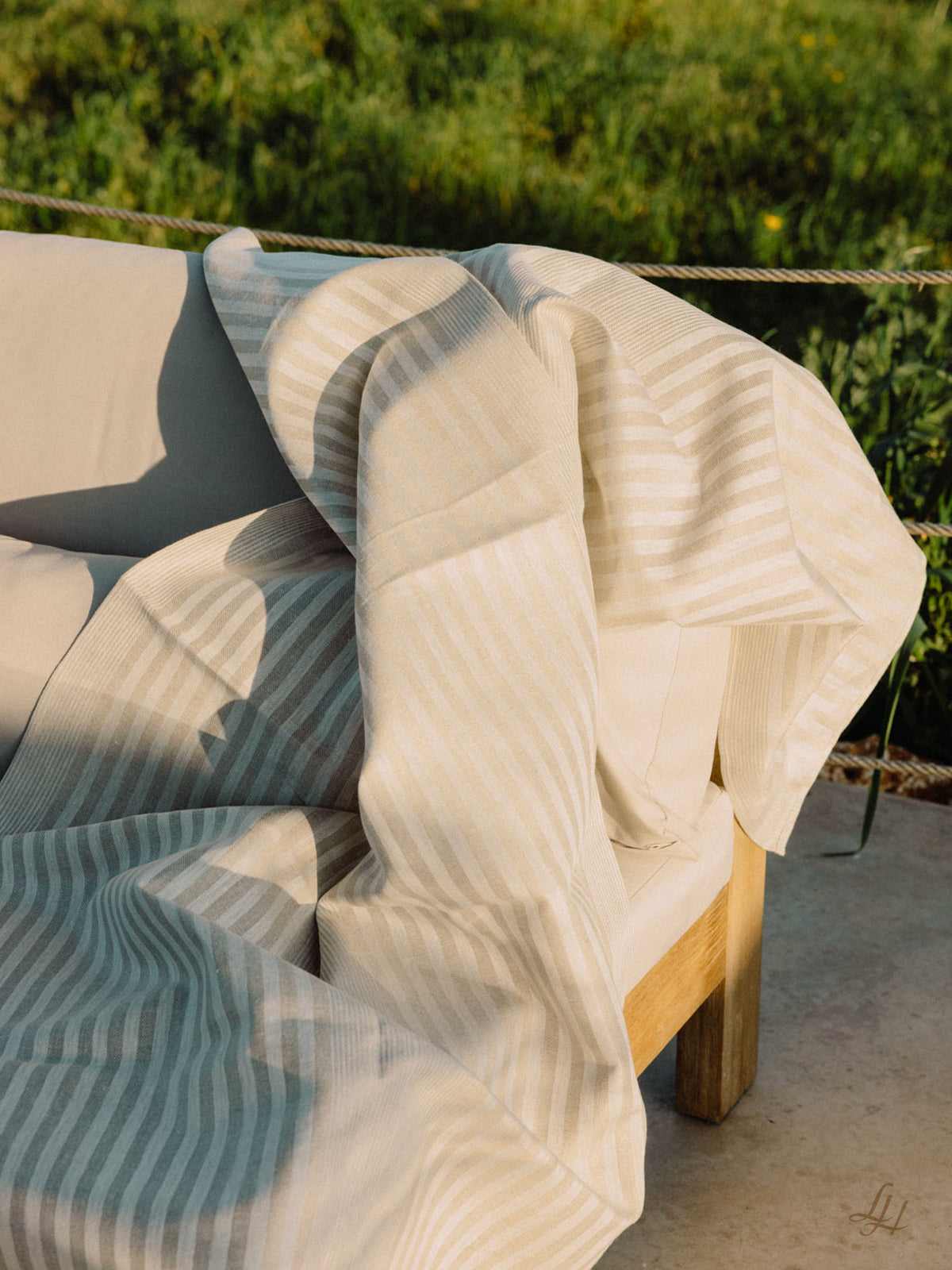 Ein beige-weißes Leinentuch, drapiert über einem hellen Sofa im Freien, mit Grünzeug im Hintergrund.