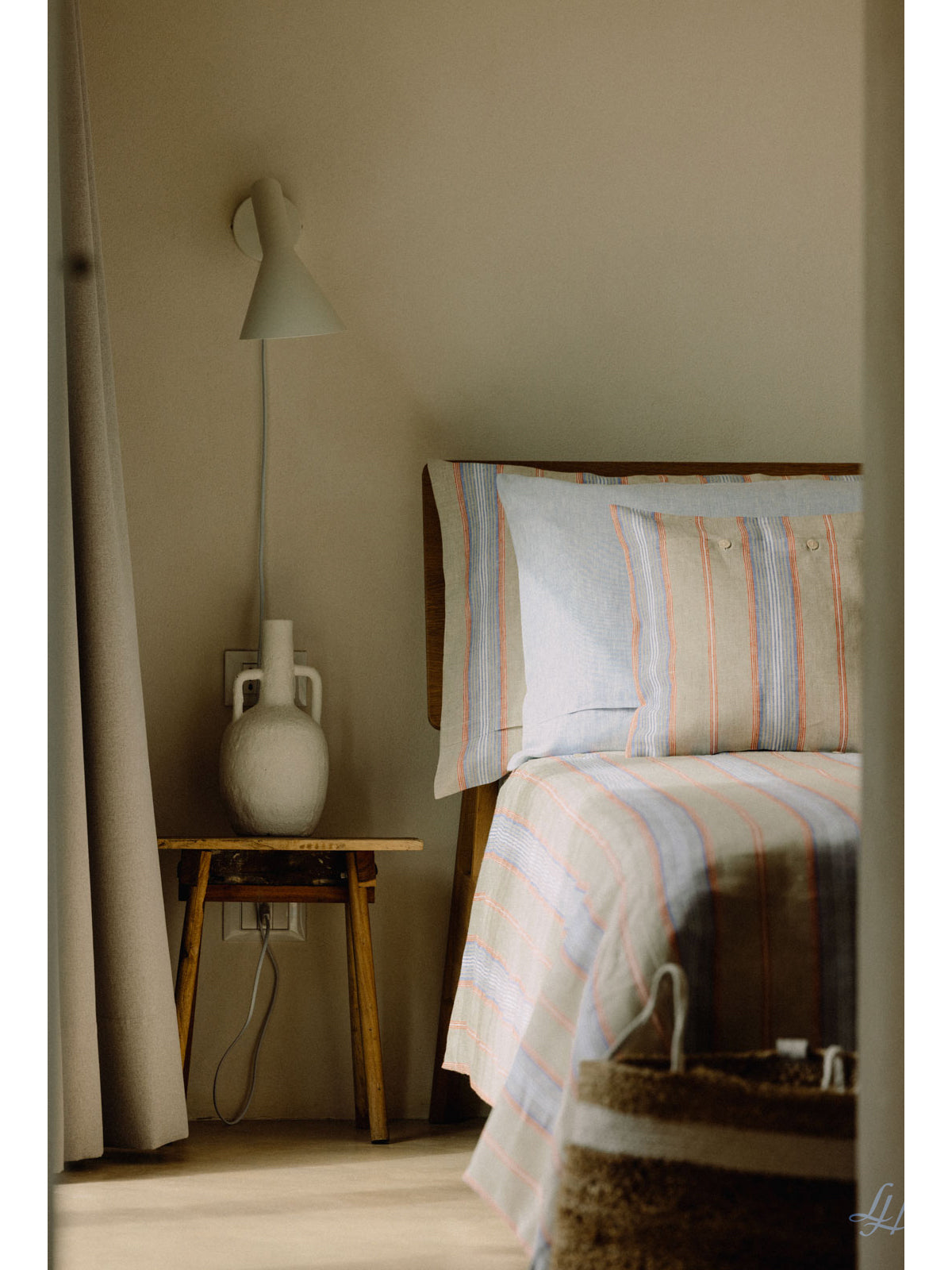Ein Bett mit gestreifter Leinenbettwäsche neben einem hölzernen Nachttisch, einer weißen Vase und einer Wandlampe in einem sanft beleuchteten Raum.