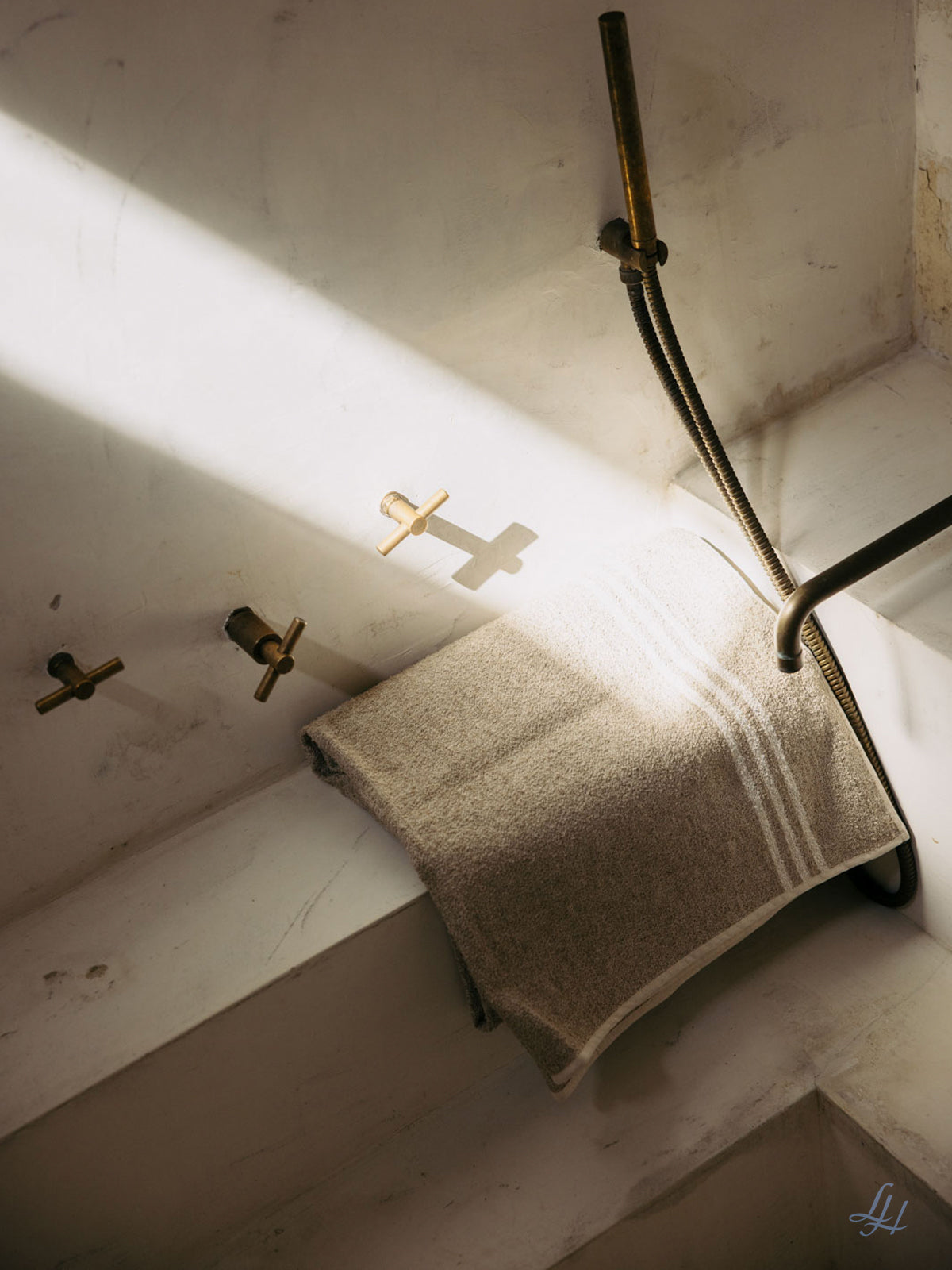 Ein beiges Leinenfrotté-Saunatuch hängt über einer weißen Badewanne neben Messingarmaturen, das Sonnenlicht wirft Schatten an die Wand.