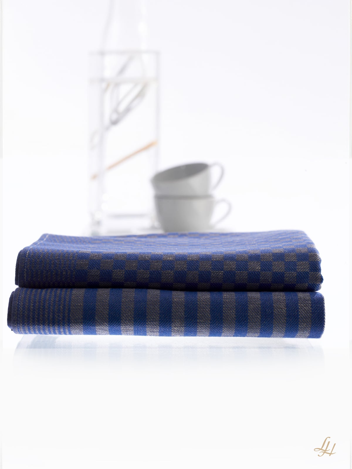 Zwei säuberlich gefaltete karierte Küchentücher liegen vor zwei weißen Tassen und einer Glaskaraffe mit Wasser gestapelt.