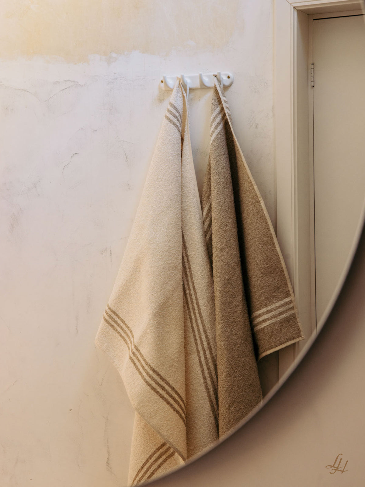 Zwei gestreifte Saunatücher aus Leinen, eines cremefarben und eines beige, hängen an Haken neben einer Tür und einem runden Spiegel an einer hellen Wand.