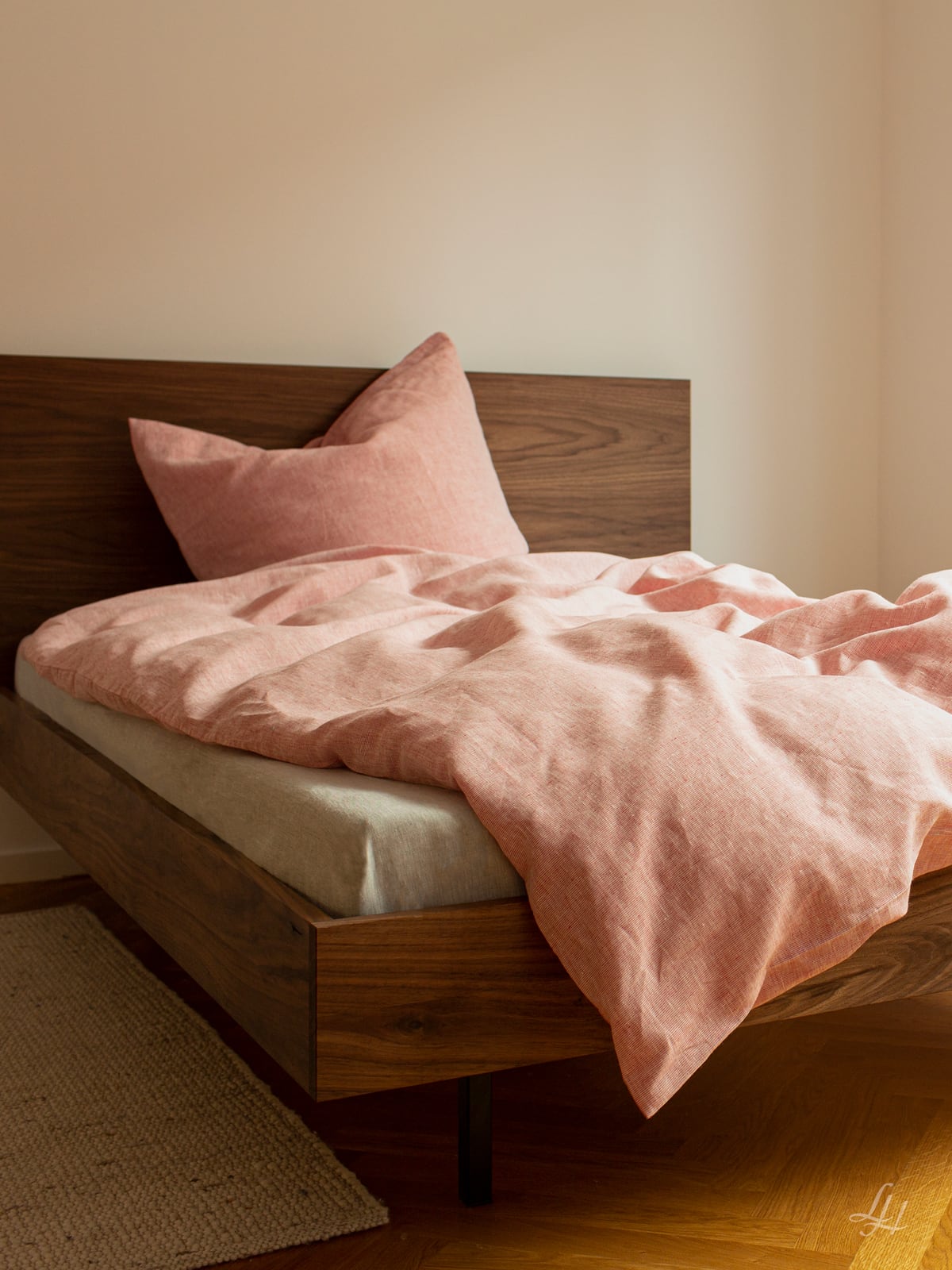 Ein Holzbett mit beiger Matratze und rosa Leinenbettbezug steht in einem minimalistisch eingerichteten Zimmer.