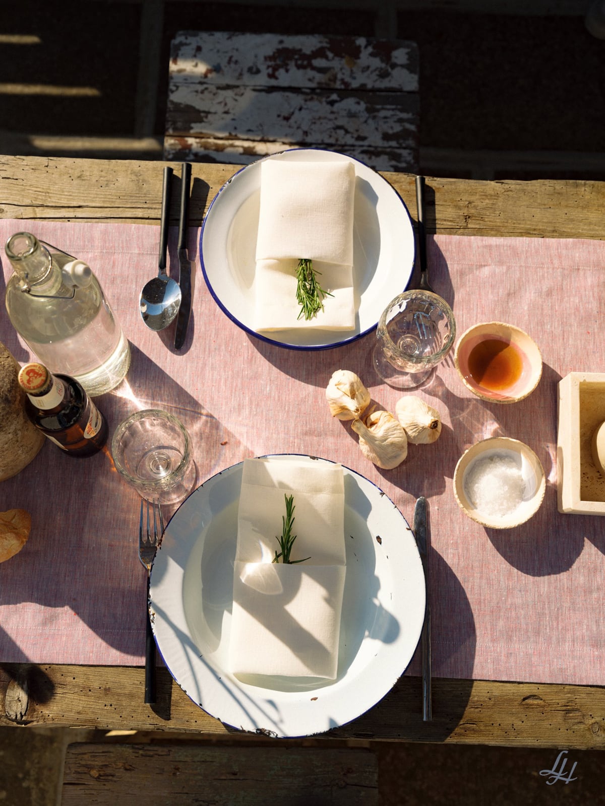 Zwei Gedecke auf einem rustikalen Tisch mit Leinen-Tischwäsche, weißen Tellern, Rosmarinzweigen, Knoblauch und erfrischenden Getränken.